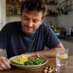 Homem brasileiro sentado à mesa de cozinha com prato de refeição sem carboidrato contendo ovos mexidos, abacate, frango grelhado e rúcula, com castanhas e copo de água ao lado
